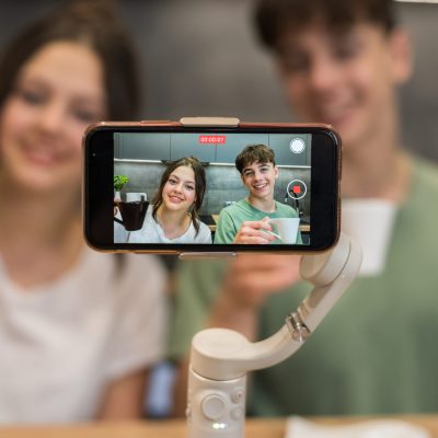 Young smiling man and woman blogger influencers talking and shooting themselves on smartphone on a stabiliser in a kitchen